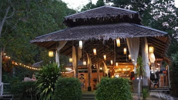 A traditional thatched-roof structure with a covered entrance, surrounded by greenery and a pathway leading to it.