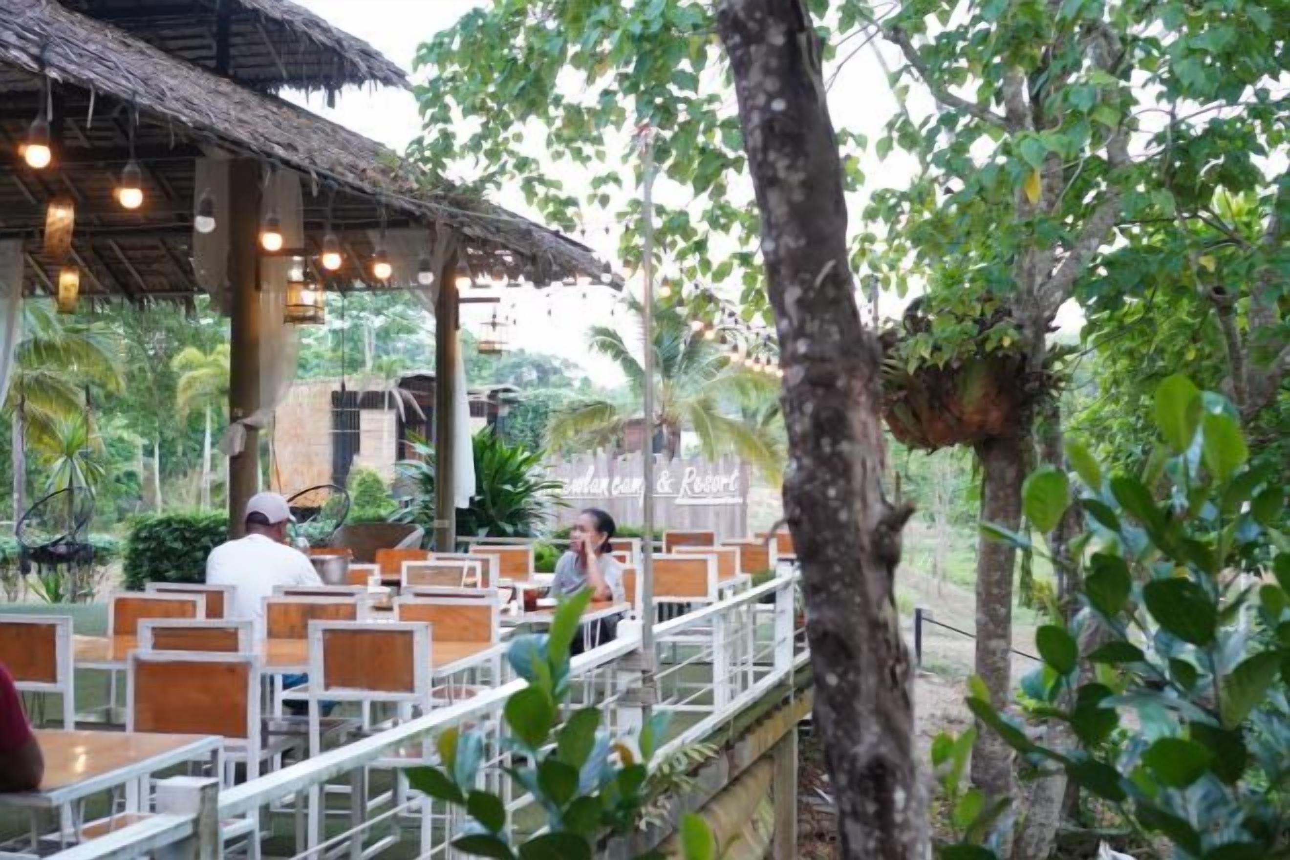 An outdoor dining area with wooden tables and chairs, surrounded by greenery and trees.