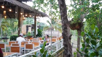 An outdoor dining area with wooden tables and chairs, surrounded by greenery and trees.