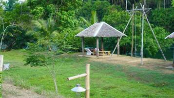 A grassy area with a thatched-roof structure, a swing set, and a small table with chairs.
