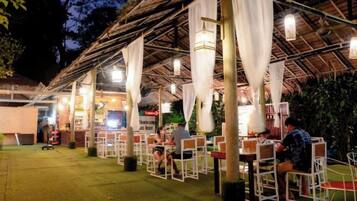 An outdoor dining area with tables and chairs, covered by a thatched roof with hanging lights.