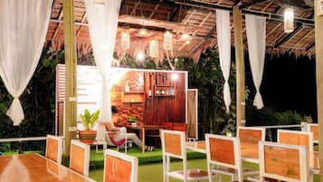 An outdoor dining area with wooden tables and chairs, a brick wall, and hanging lights.