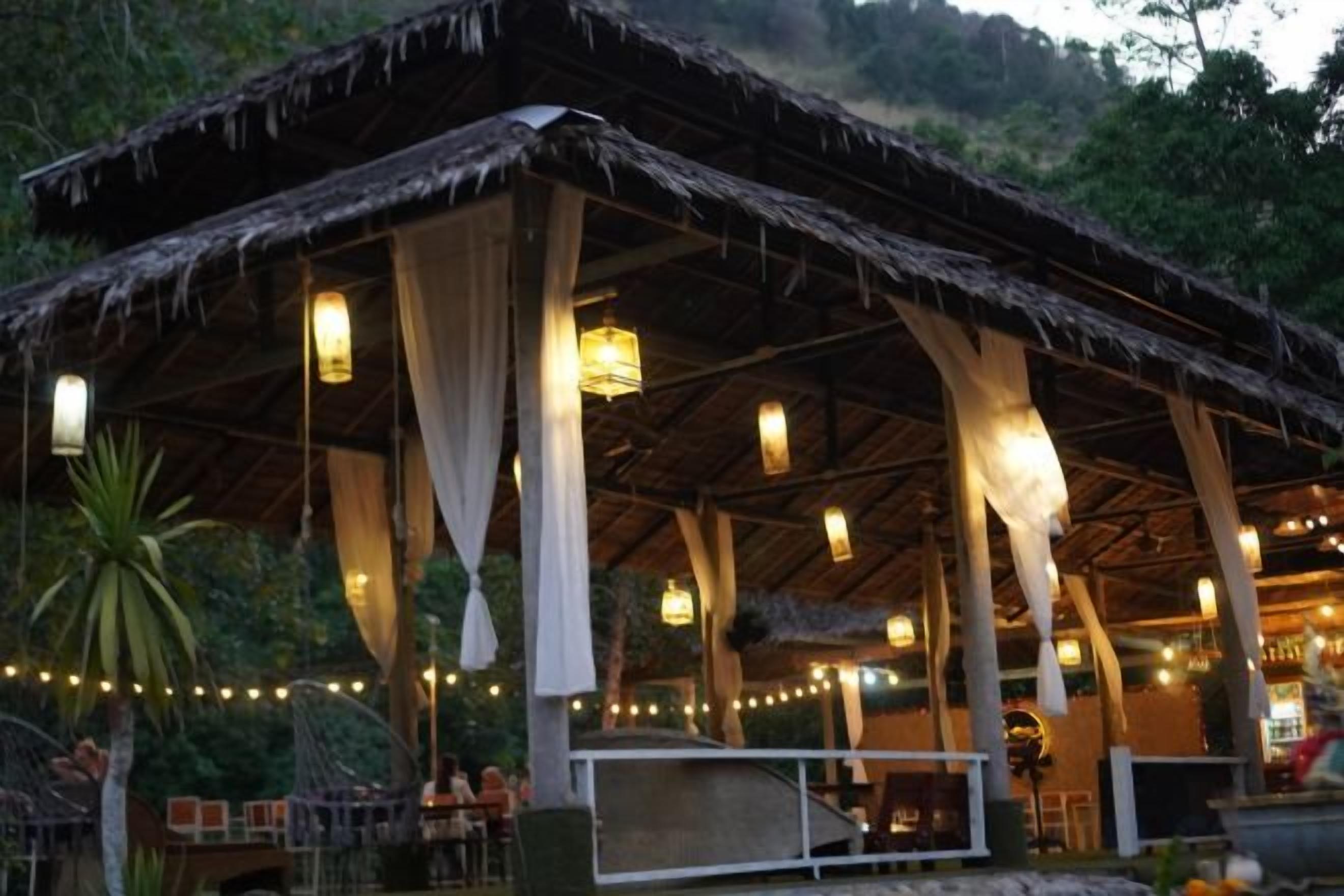 Un área de comedor al aire libre con techo de paja, luces colgantes y mesas.