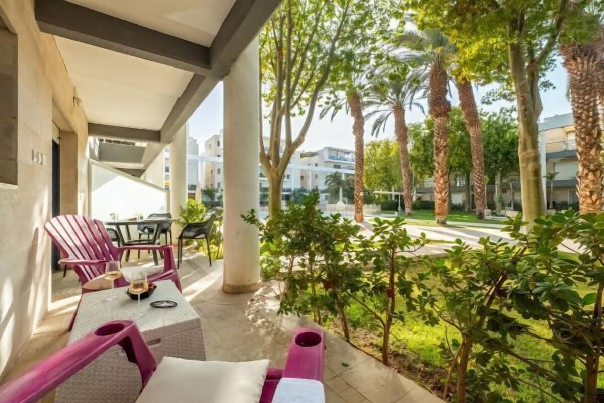 A covered outdoor seating area with chairs and a table, surrounded by greenery and palm trees.