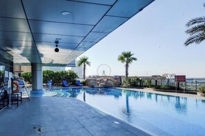 Una piscina exterior con sillones de descanso, palmeras y una noria al fondo.