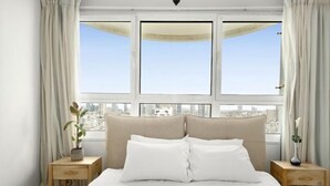 A bedroom with a large bed, two bedside tables, a potted plant, and a view of the city through large windows. - Magnificent Seaside Panorama Stay by Sea N' Rent (Tel Aviv)