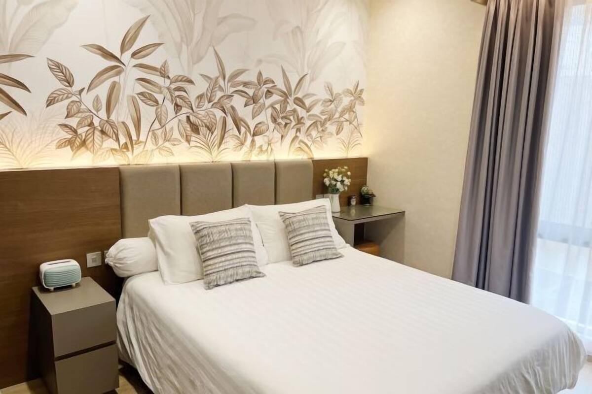 Una cama bien hecha con sábanas blancas y dos almohadas, una mesita de noche con una radio, y una pared con un diseño de hojas.