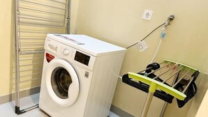 A compact, white front-loading washing machine in a small laundry area with a metal drying rack and a wall-mounted electrical outlet. - Premium Luxury Home At Golf Island PIK (North Jakarta)