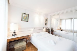 Una habitación de hotel con dos camas, un escritorio con lámpara, un pequeño banco y un espejo.