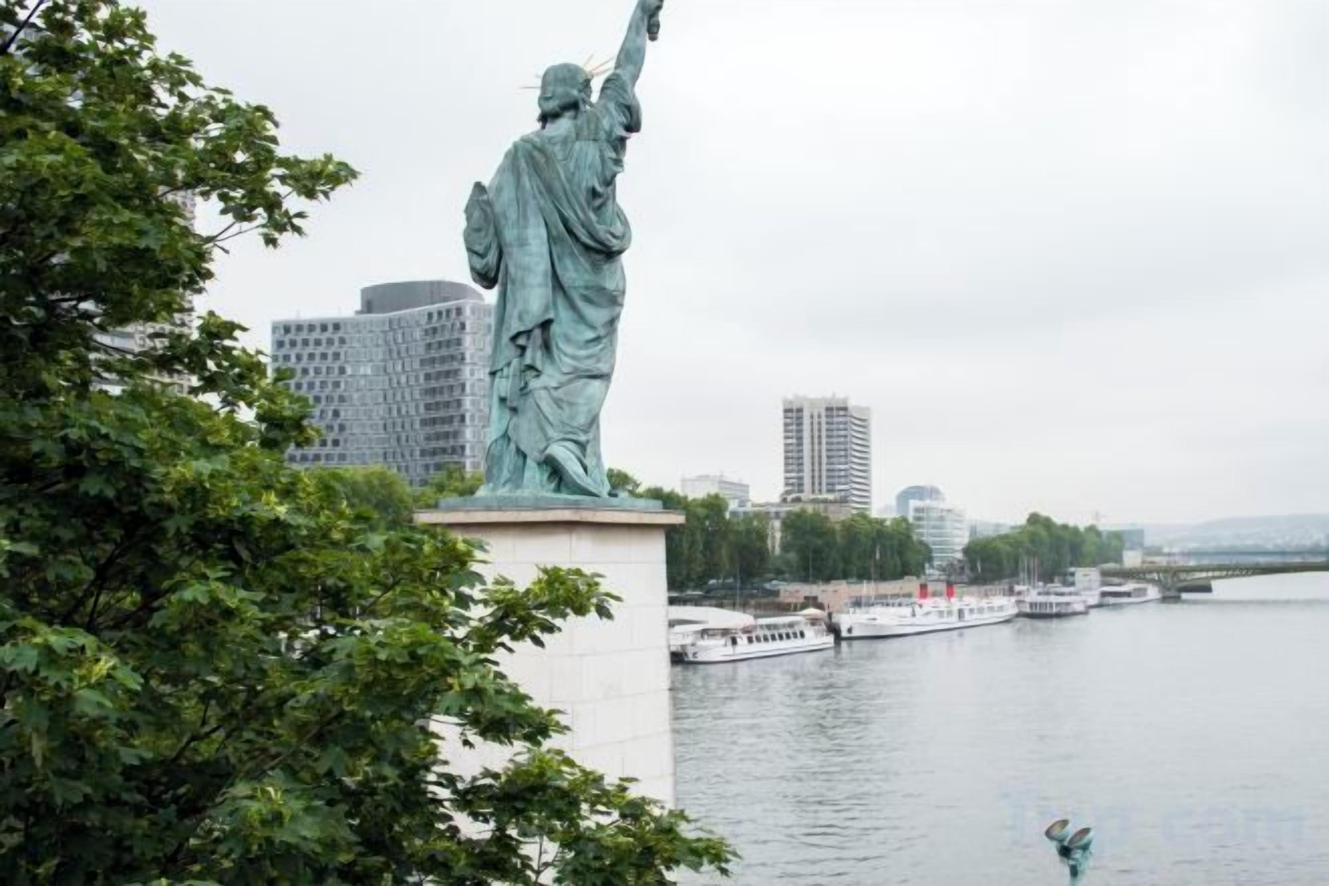 A statue of a figure holding a torch, with a body of water and buildings in the background.