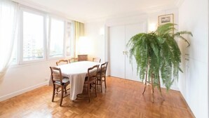 A dining room with a round table and chairs, large windows, and a large green plant.