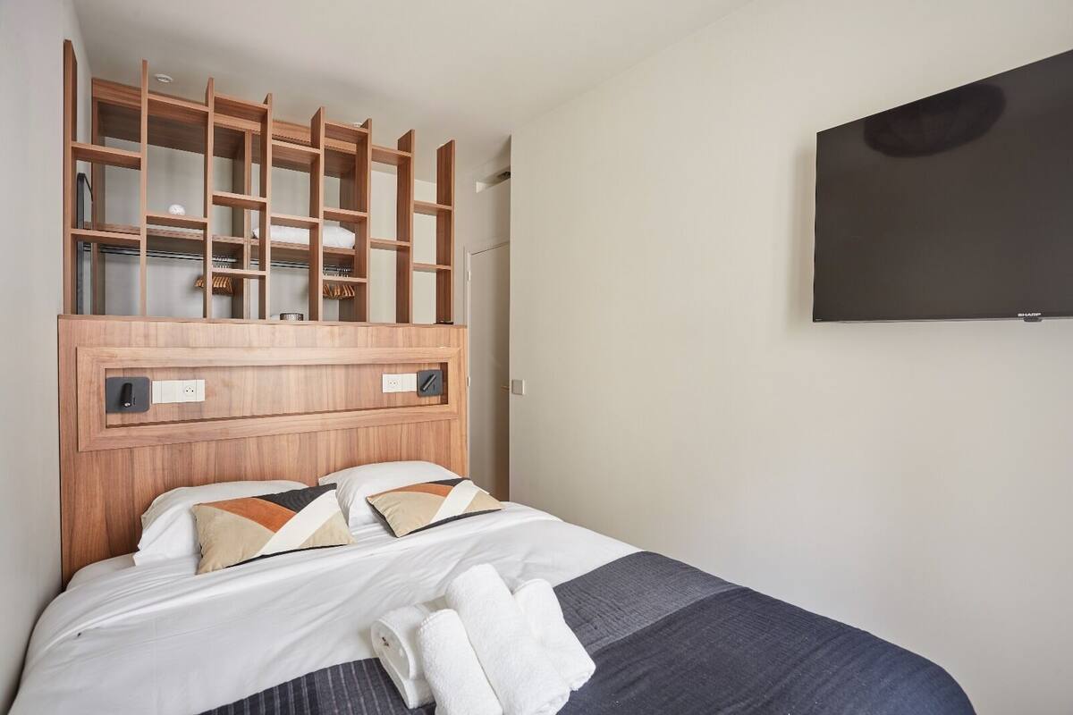 A hotel room with a wooden headboard, a bed with white and patterned pillows, a flat-screen TV mounted on the wall, and a neatly made bed with towels.