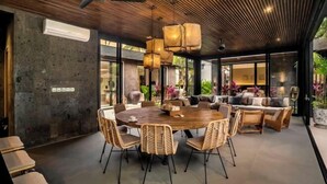 Un espace repas moderne avec une table ronde en bois et des chaises en osier, entouré de canapés et d’une grande fenêtre donnant sur un paysage verdoyant.