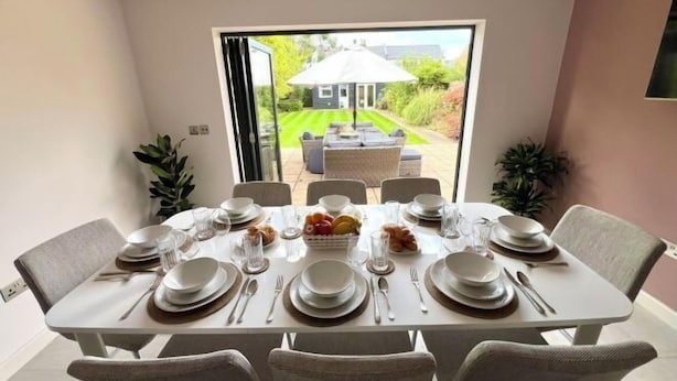 A dining room with a table set for six, a garden view through a glass door, and a patio area with outdoor furniture. - Luxury Family Home in Central Windsor with Large Garden, Games Room & Parking (Windsor)