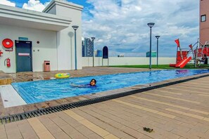 Une piscine où un enfant nage, entourée d’un bâtiment doté d’une porte, d’un extincteur et d’un toboggan rouge.