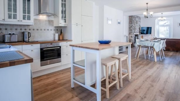 Une cuisine moderne avec des meubles blancs, un îlot central doté de tabourets, un coin repas avec une table et des chaises, et une cheminée en pierre.