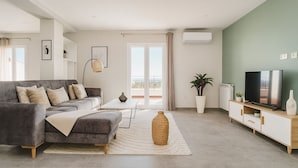 A modern living room with a grey sofa, a white coffee table, a TV on a wooden stand, and a large window with a view of the outdoors. - Spacieux Appartement a Dely Brahim (Algiers)