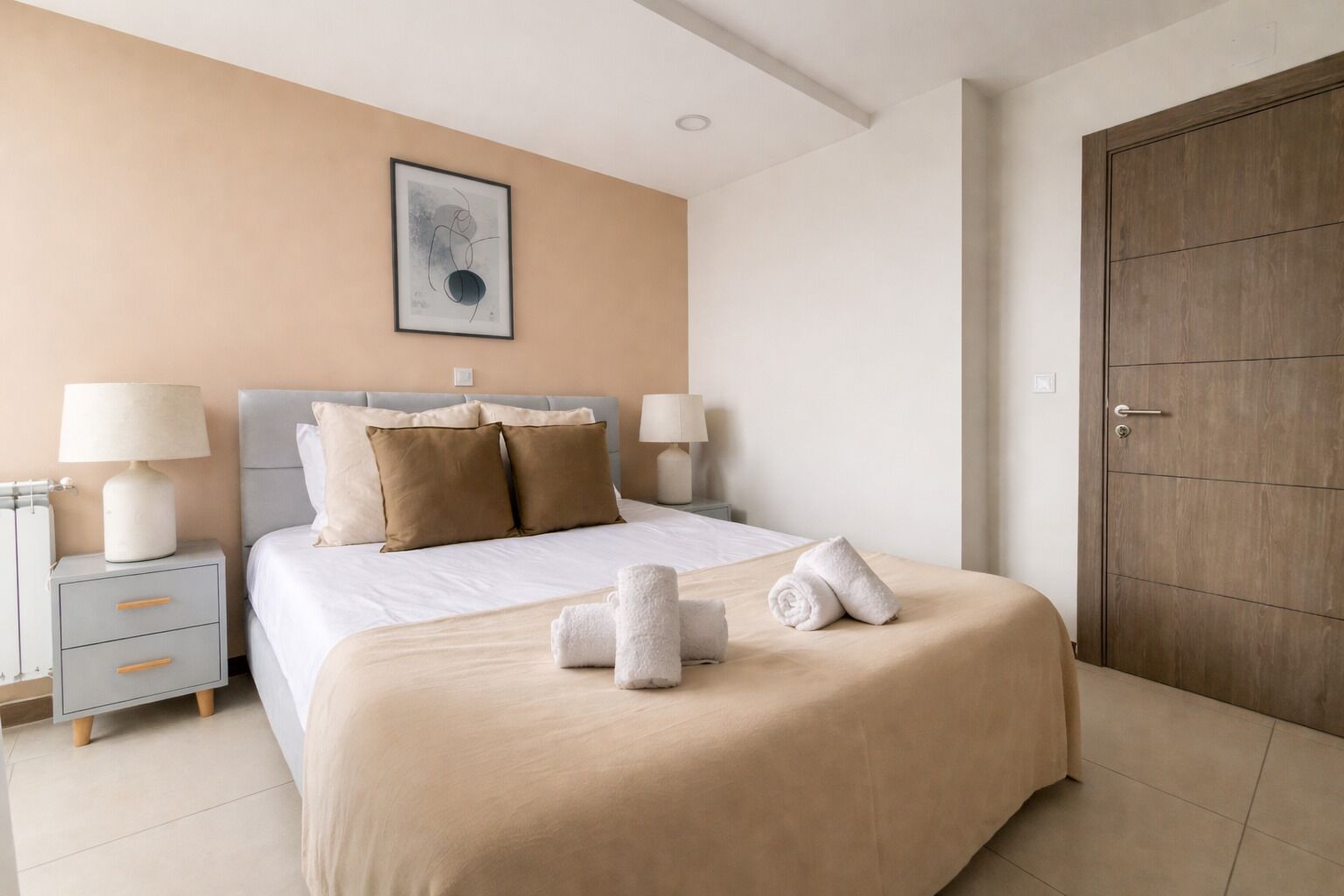 Un cuarto de hotel ordenado con cama, lámparas de noche, una obra de arte enmarcada y una puerta de madera.