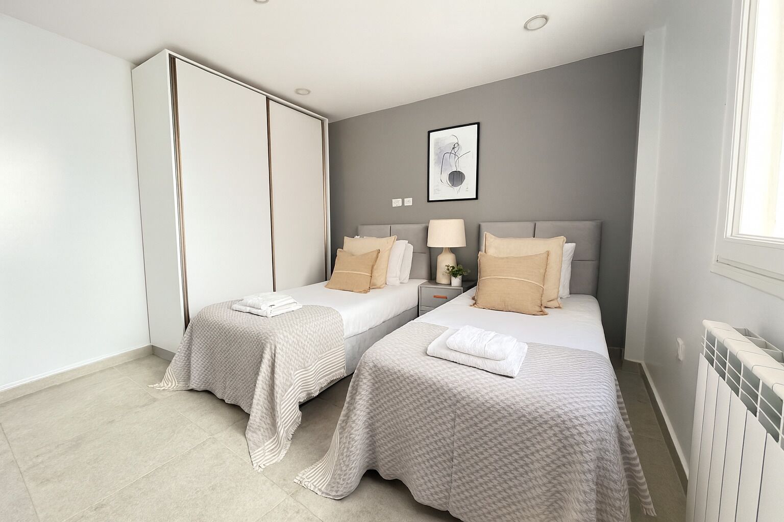 A modern bedroom with two beds, a wardrobe, a bedside table with a lamp, and a framed picture on the wall.