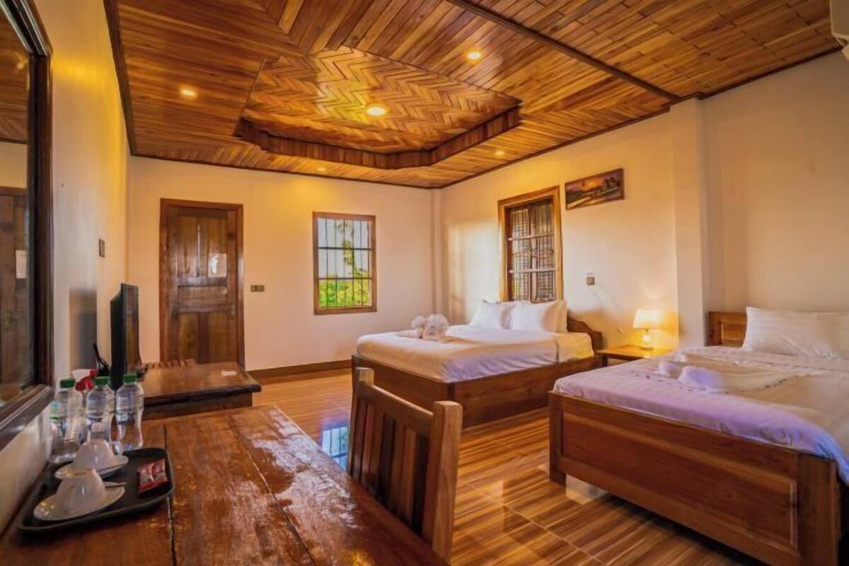 Une chambre d’hôtel avec deux lits en bois, une table à manger en bois sur laquelle se trouve un plateau avec des bouteilles d’eau, et un téléviseur.