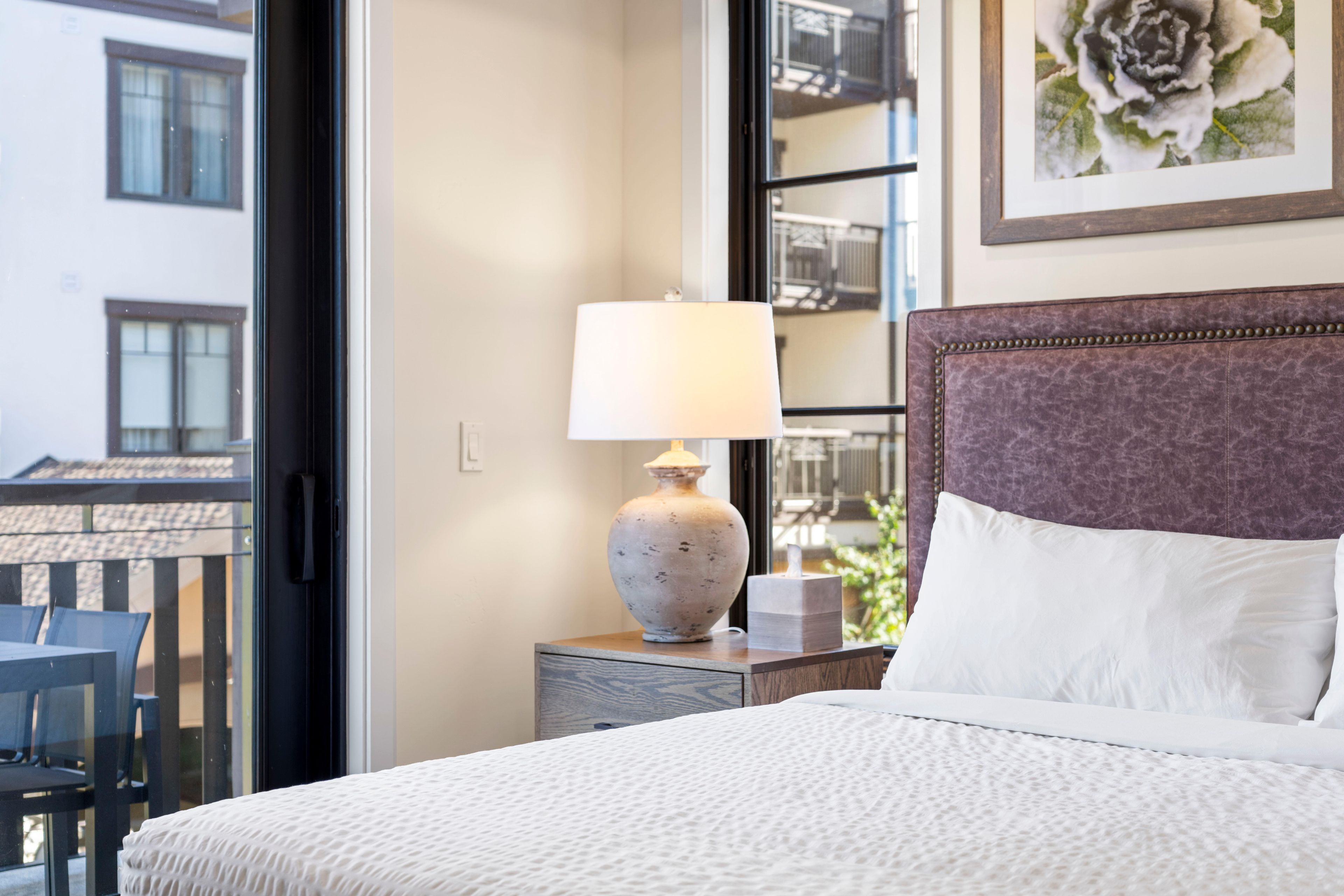 Une chambre d’hôtel avec un lit, une table de chevet avec une lampe, une chaise et une fenêtre donnant sur des immeubles.