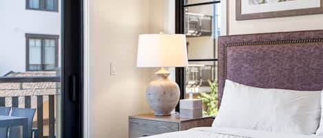 A hotel room with a bed, a nightstand with a lamp, a chair, and a window with a view of buildings.