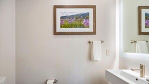 A bathroom with a framed picture of flowers on the wall, a towel rack with a towel, a toilet paper holder, and a sink with a faucet. - Snowcation Station | Exclusive Tamarack Perks (Tamarack)