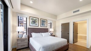 A modern bedroom with a large bed, two bedside tables with lamps, a wooden headboard, and a view of the outside through a window. - Snowcation Station | Exclusive Tamarack Perks (Tamarack)