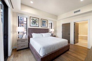 Une chambre moderne avec un grand lit, deux tables de chevet avec des lampes, une tête de lit en bois et une vue sur l’extérieur par une fenêtre.