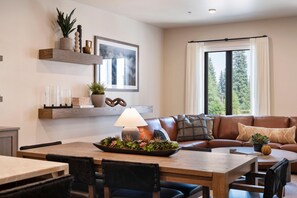 A modern living room with a dining area, featuring a brown leather sofa, a wooden dining table, and a large window with curtains.