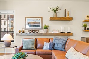 Een woonkamer met een bruine leren bank, een lijst met een landschapsafbeelding en houten planken met decoratieve objecten.