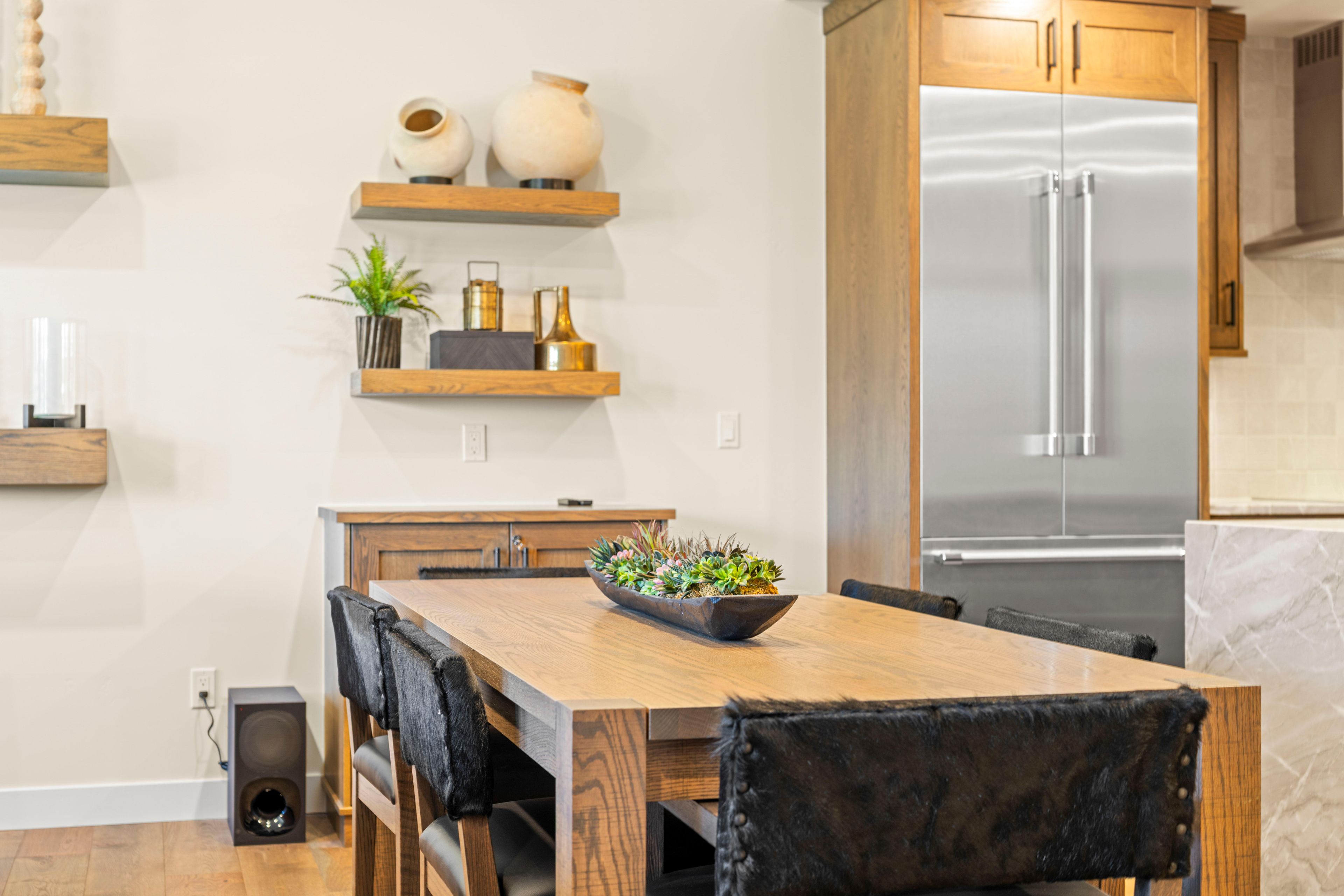 Une cuisine moderne avec une table à manger en bois, des chaises, un réfrigérateur en acier inoxydable encastré et des objets décoratifs disposés sur des étagères.