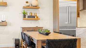 Une cuisine moderne avec une table à manger en bois, des chaises, un réfrigérateur en acier inoxydable encastré et des objets décoratifs disposés sur des étagères.