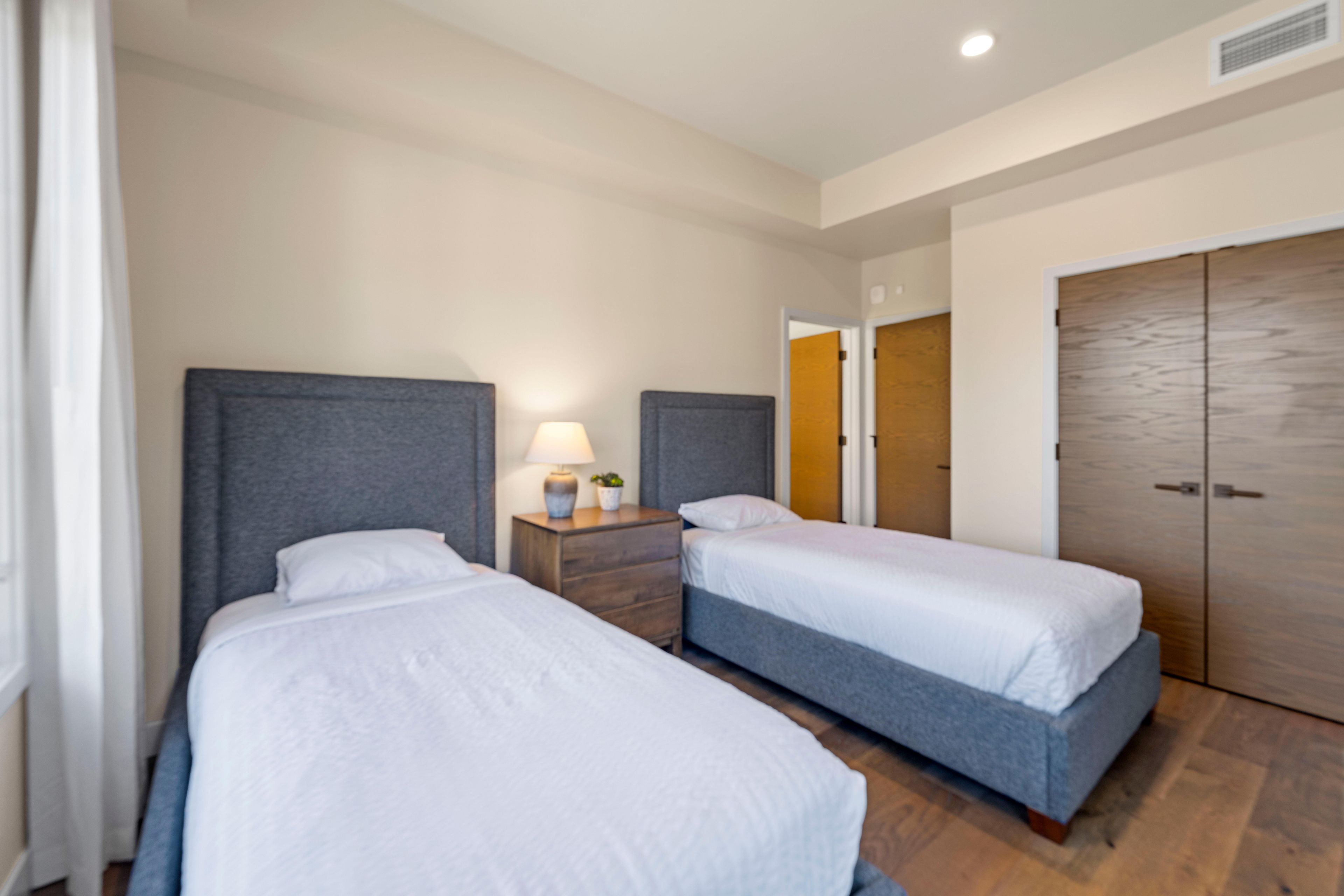 Habitación de hotel con dos camas individuales, una mesita de noche con lámpara, un armario y una puerta que da a otra habitación.