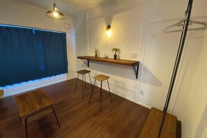 A room with a wooden floor, a hanging light fixture, a shelf with plants, and a window with a curtain. - Poroto Base (Shiraoi)