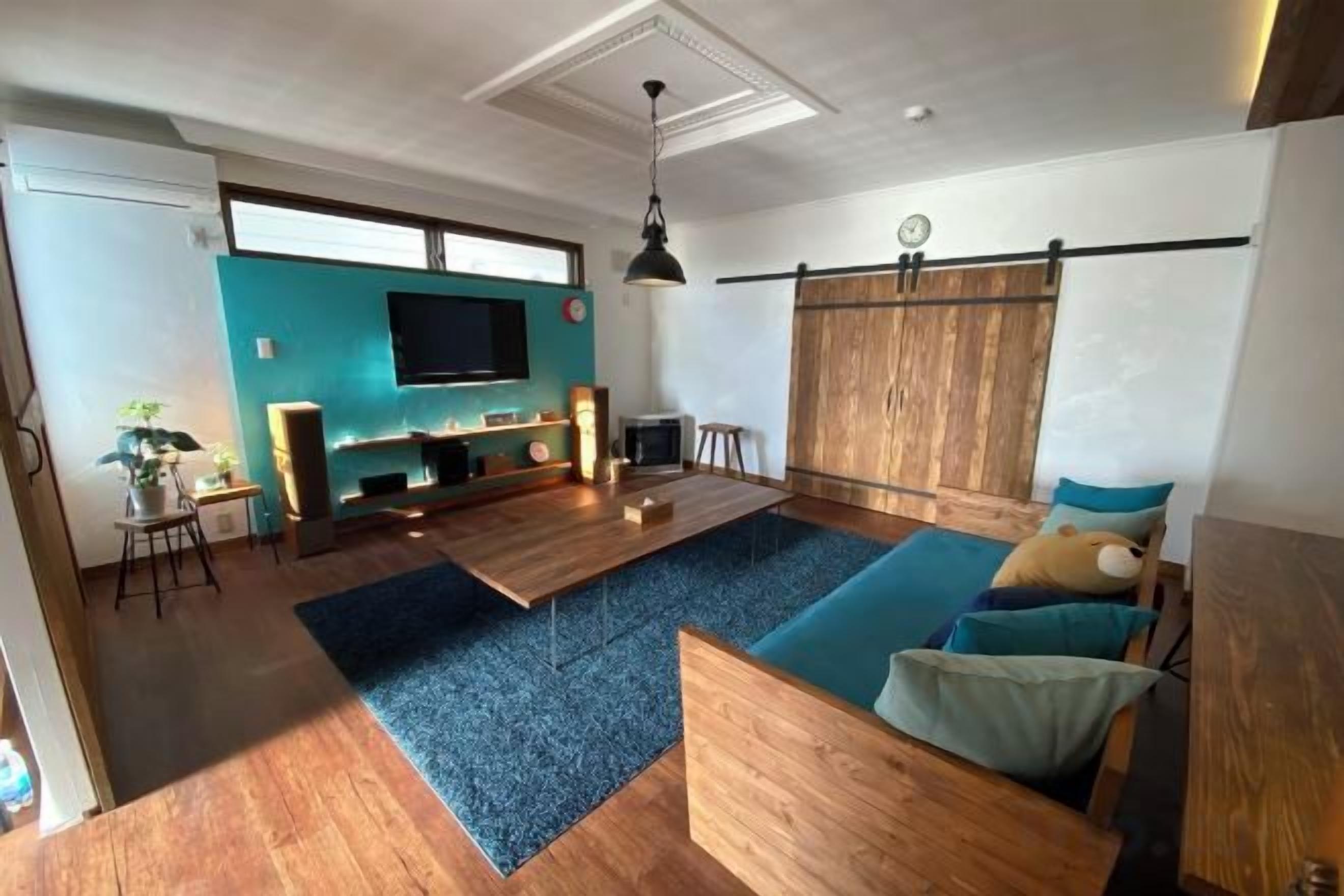 A living room with a wooden floor, a blue carpet, a sofa with cushions, a TV mounted on the wall, and a wooden partition.