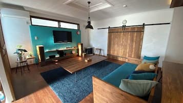 A living room with a wooden floor, a blue carpet, a sofa with cushions, a TV mounted on the wall, and a wooden partition.