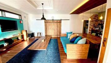 A modern living room with a large TV, wooden furniture, a blue carpet, and a kitchen area with hanging lights.