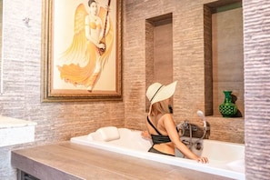 A woman in a bathtub with a towel, a framed painting on the wall, and a green vase on a shelf.
