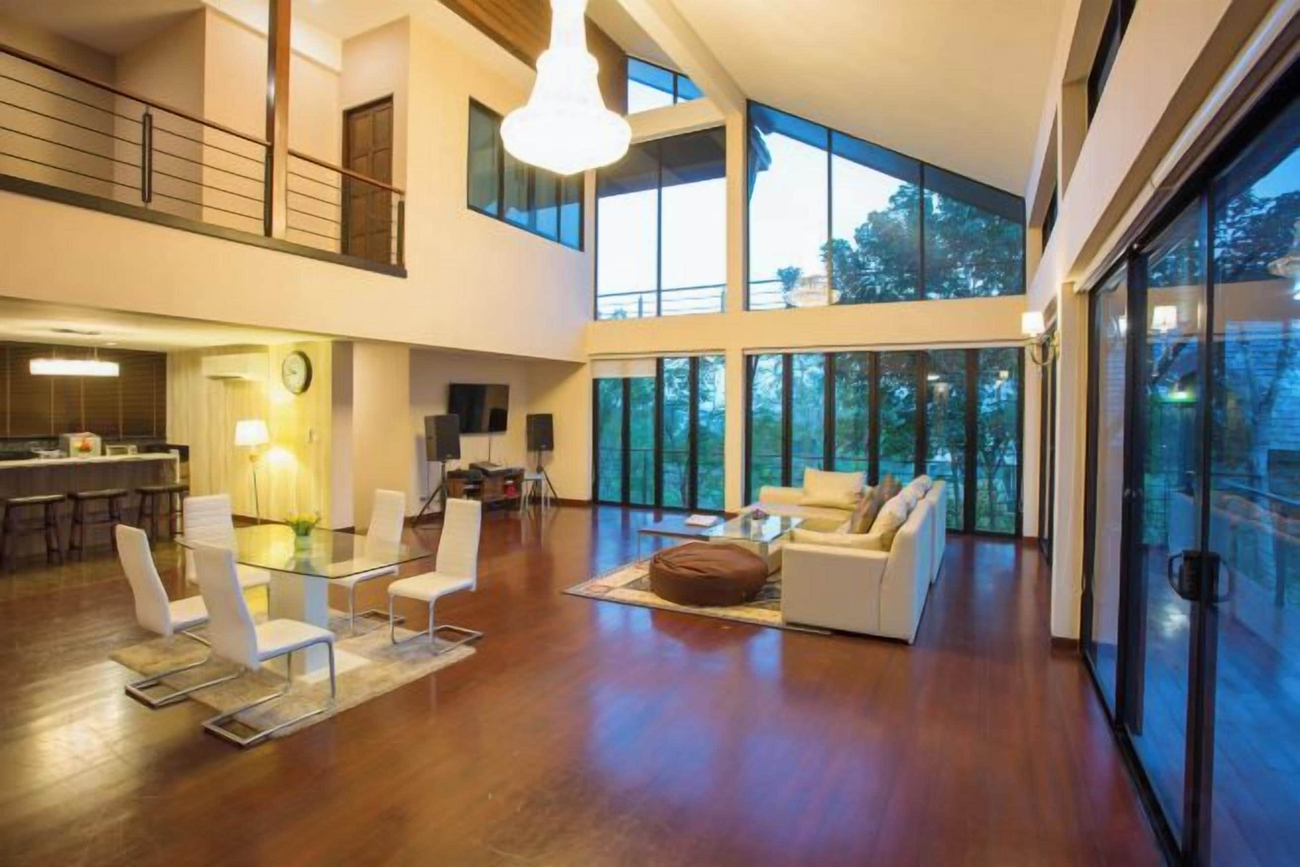 A spacious living area with a high ceiling, a dining area with modern furniture, and large windows offering views of the outdoors.