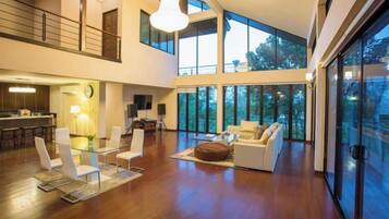 A spacious living area with a high ceiling, a dining area with modern furniture, and large windows offering views of the outdoors.