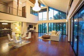 A spacious living area with a high ceiling, a dining area with modern furniture, and large windows offering views of the outdoors. - Naralin Khaoyai (Pak Chong)