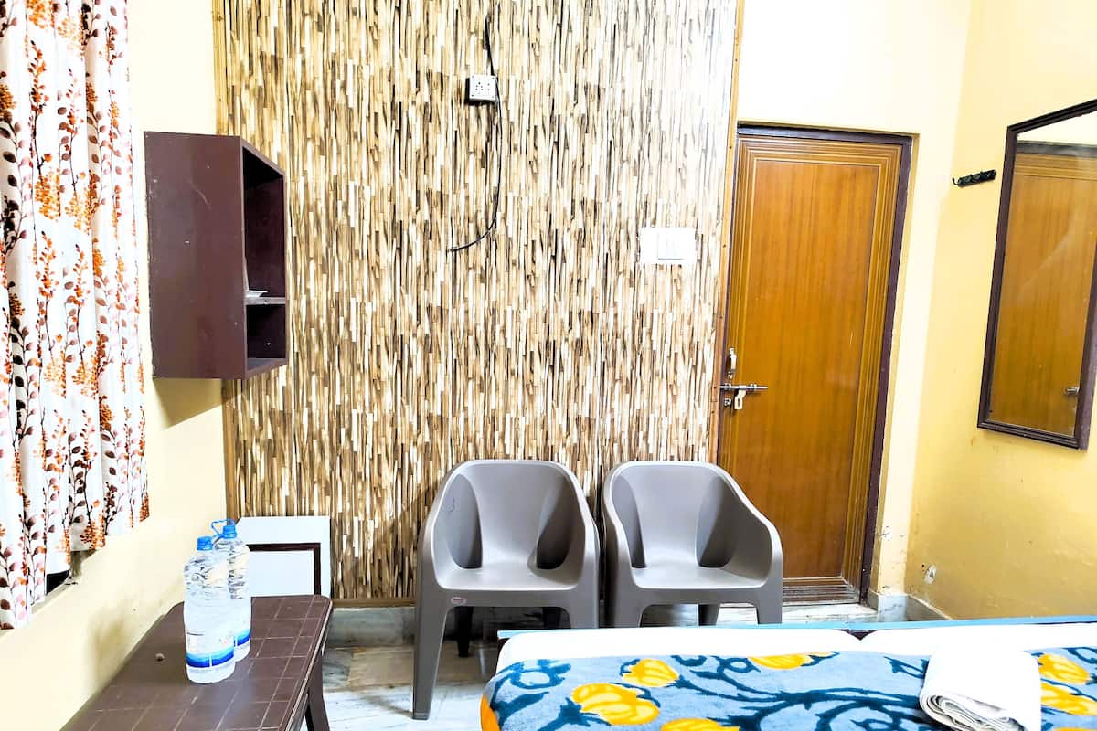 A hotel room with a bed, two chairs, a small table with water bottles, a wall-mounted shelf, and a wooden door.