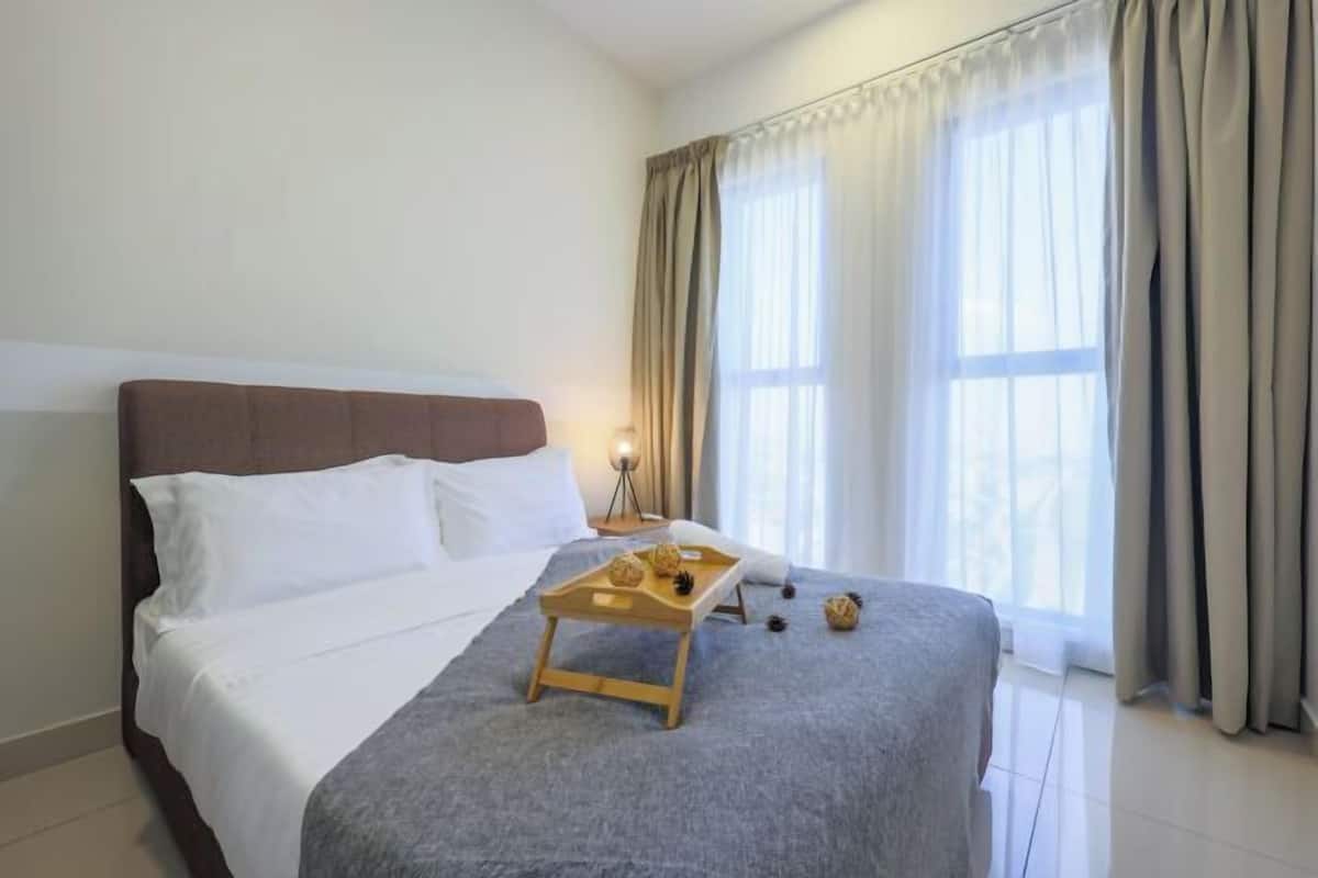 Una cama bien hecha con una bandeja de aperitivos, una lámpara de noche y una ventana con cortinas.