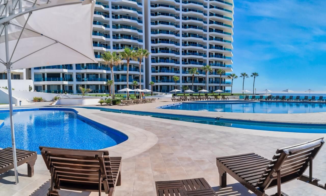 Ein Hotelpoolbereich mit mehreren Schwimmbecken, Liegestühlen und einem großen Gebäude im Hintergrund.