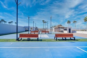 A blue tennis court with wooden benches, palm trees, and a thatched-roof structure.