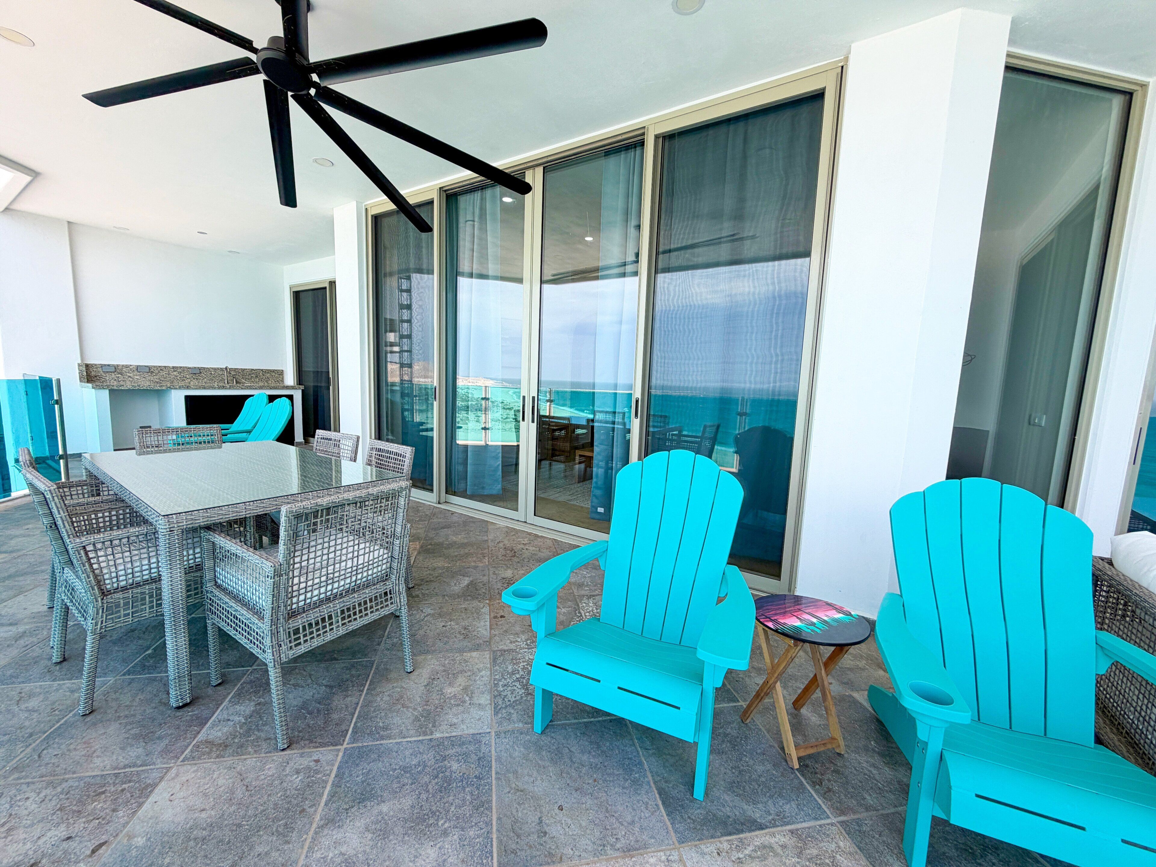 A sunlit patio with a glass door leading to a balcony, a table with chairs, and a ceiling fan.