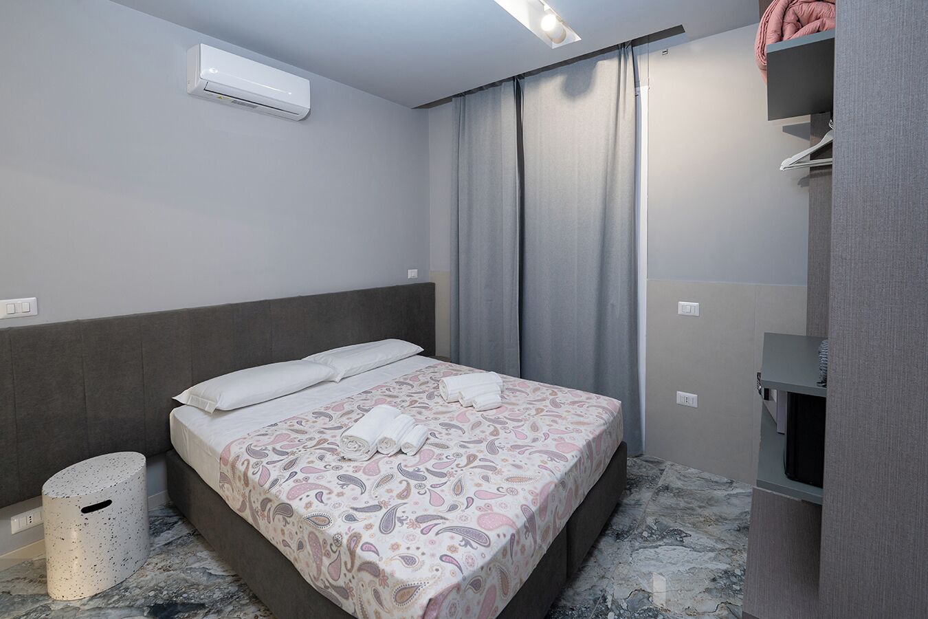 A hotel room with a bed, a wardrobe, a trash bin, and an air conditioning unit.