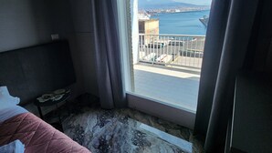 A hotel room with a bed, a small table, and a view of a port with boats and a distant mountain. - Mia Suites Angioino (Naples)