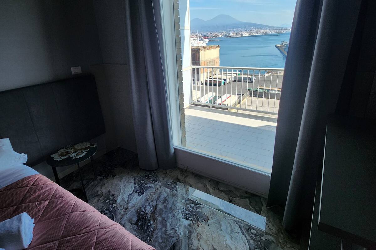 Habitación de hotel con cama, una mesita y vistas a un puerto con barcos y una montaña distante.